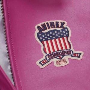 Pode incluir: Close-up de um artigo de couro magenta com z&iacute;per. Uma aplica&ccedil;&atilde;o bordada &eacute; costurada na frente, apresentando um design de escudo com o texto "AVIREX ESTABLISHED NY 1975" e um esquema de cores vermelho, branco e azul.