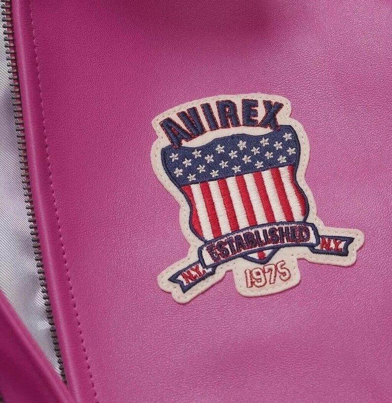 Pode incluir: Close-up de um artigo de couro magenta com z&iacute;per. Uma aplica&ccedil;&atilde;o bordada &eacute; costurada na frente, apresentando um design de escudo com o texto "AVIREX ESTABLISHED NY 1975" e um esquema de cores vermelho, branco e azul.