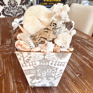 May include: A decorative square vase with a gray and white Asian-inspired design, filled with seashells, coral, and driftwood. The vase has a gold rim and sits on a wooden table. The arrangement includes various textures and natural elements.