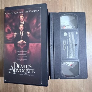 May include: A black VHS cassette tape and its matching case for the film Devil's Advocate. The case features Keanu Reeves and Al Pacino against a dark red background with the movie title and text about the plot. The tape has a black plastic shell.