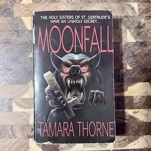 May include: A paperback book titled Moonfall by Tamara Thorne with a black cover featuring a snarling, dark-furred creature with glowing pink eyes and sharp teeth, holding an inverted cross. The text reads: The holy sisters of St. Gertrude's have an unholy secret.