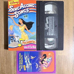 May include: A Disney Sing Along Songs VHS tape featuring Pocahontas and the song "Colors of the Wind." The box is orange with illustrations of Pocahontas, a raccoon, and other characters. Also included is a VHS tape and a purple checklist.