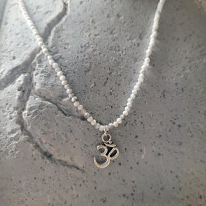 Könnte beinhalten: Weißes Perlenhalskette mit einem silberfarbenen Om-Anhänger. Die Halskette besteht aus kleinen, runden weißen Perlen, die aneinandergereiht sind. Der Om-Anhänger ist ein stilisiertes Symbol mit dunklem Akzent. Die Halskette wird vor einem strukturierten grauen Hintergrund präsentiert.
