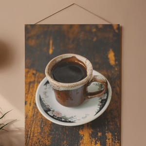 Puede incluir: Una taza de café de cerámica marrón llena de líquido oscuro, sobre un platillo a juego. La taza tiene un borde texturizado y un asa. El platillo presenta un diseño floral en el borde. La imagen está montada en un panel de madera.