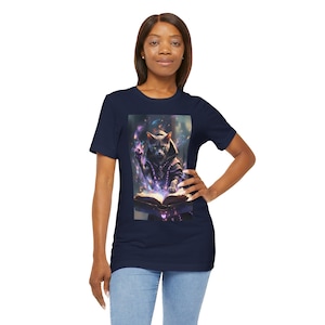 May include: Navy blue t-shirt featuring a fantasy illustration of a cat wearing a hooded robe, reading a glowing book. The image has a cosmic background with purple and white accents. The cat is the central focus of the design.