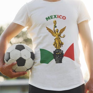 Puede incluir: Camiseta blanca con un gráfico de una estatua dorada sosteniendo un trofeo, la palabra "MEXICO" en verde y rojo, y un balón de fútbol. La camiseta también presenta los colores de la bandera mexicana.