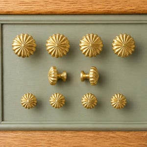 Peut inclure: Une collection de dix boutons de placard en métal doré disposés en rangées. Les boutons présentent un design circulaire, cannelé ou festonné avec des lignes rayonnantes partant du centre. Deux boutons sont vus de côté, révélant leurs tiges.