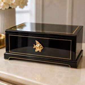 May include: A rectangular black storage box with a glossy finish and gold-toned trim. The front features a centred gold-toned metal ornament shaped like an angelfish, which serves as a handle for the drawer. The box sits on a raised base.