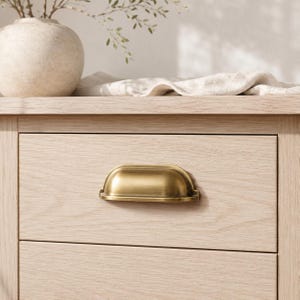 May include: Close-up of a light wood dresser drawer with a brass cup pull. The drawer is part of a larger wooden cabinet. A vase with greenery and a linen cloth are on top of the cabinet.