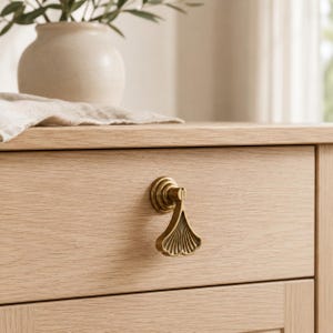 Puede incluir: Primer plano de un armario de madera clara con un tirador de cajón de color latón. El tirador del cajón tiene forma de abanico u hoja de ginkgo. El armario tiene un acabado de veta de madera clara. Un jarrón con vegetación está en el fondo.