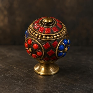 Peut inclure: Un bouton de meuble sphérique en laiton avec une base en métal sombre incrustée d'accents en pierre rouge et bleue. Le design comprend un anneau central en laiton perlé, des motifs géométriques en tuiles rouges et des motifs floraux.