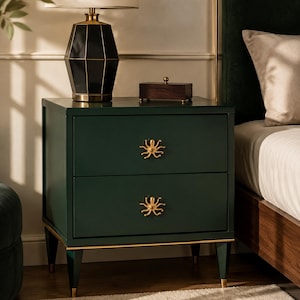 May include: A dark green bedside table featuring two drawers with gold-toned octopus-shaped drawer pulls. The table is supported by four tapered legs with gold-toned feet. The surface holds a black geometric lamp and a small wooden box.