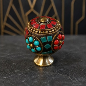 May include: A spherical cabinet knob featuring a dark brown base with inlaid red and turquoise stone accents. The knob is decorated with small brass beads and floral patterns, finished with a polished brass pedestal base and a central brass dome.