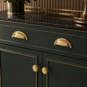 May include: A dark green cabinet featuring a black marble top with white veining. The cabinet includes two drawers with polished gold cup pulls and two doors with matching round gold knobs, all accented by thin gold trim along the edges.