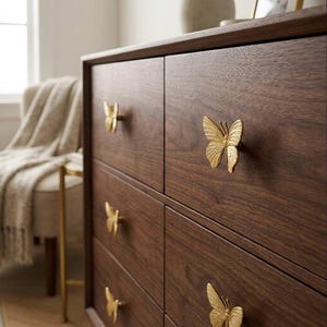 May include: A dark brown wooden dresser with six drawers, each adorned with a gold butterfly-shaped handle. The butterfly handles add a decorative touch to the otherwise simple design. The dresser is a piece of bedroom furniture.