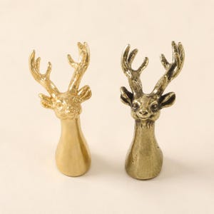 May include: Two deer head figurines crafted from metal. The left figurine features a smooth, polished gold-tone finish, while the right figurine displays a textured, antique brass-tone finish. Both items depict a stag with detailed antlers.