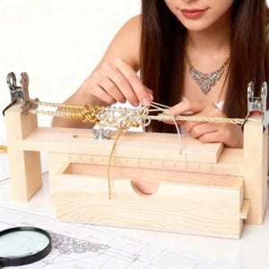 May include: A light-coloured wooden jig for crafting paracord or rope bracelets, featuring a built-in ruler with markings in centimetres, a storage drawer, two metal clips at the ends, and a central wing nut for adjusting the length of the cord project.