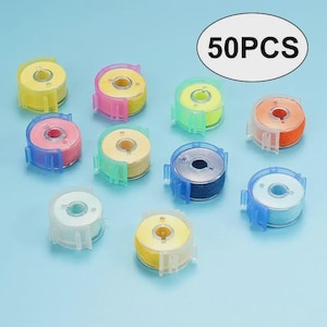 May include: A collection of 50 sewing machine bobbins in various colors, including yellow, pink, green, orange, blue, and white. Each bobbin is filled with thread and encased in a clear plastic housing. The bobbins are arranged on a light blue surface.