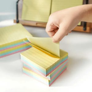 May include: A hand pulling a yellow lined sticky note from a stack of colorful sticky notes. The edges of the stack show layers of pink, blue, and yellow. Additional stacks of sticky notes are visible in the background.