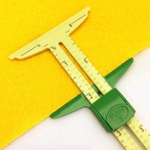 May include: A yellow and green plastic sewing gauge with a sliding marker. The tool features imperial markings in inches, including fractions like 1/4 and 1/2. A green sliding component with the word PUSH embossed on it allows for adjustment.
