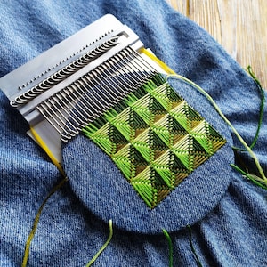 May include: A denim patch featuring a geometric green and brown embroidered design. A metal weaving tool is placed over the patch, with strands of green and yellow thread extending from the design.