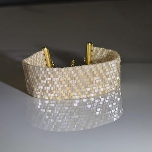 Op de afbeelding: Een brede armband van geweven kralen met een herhalend geometrisch patroon in afwisselende tinten iriserend wit en zacht beige. De armband is afgewerkt met twee goudkleurige cilindrische metalen eindkappen die de kralenband vastzetten.
