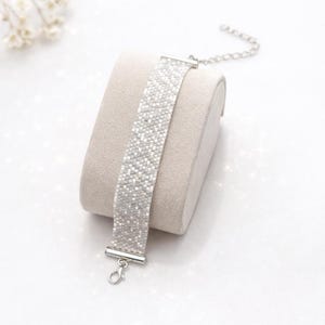 May include: A wide bracelet crafted from small, shimmering white and silver beads arranged in a woven pattern. The bracelet features silver-toned metal end caps, a lobster claw clasp, and an adjustable chain extension for a secure fit.
