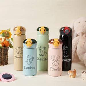 Peut inclure: Cinq bouteilles d'eau cylindriques de couleur beige, bleu clair, vert sauge, rose clair et noir. Chaque bouteille est dotée d'un couvercle à rabat avec un loquet jaune ou rouge, d'une illustration d'animal et d'un prénom imprimé en police serif.