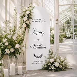 Può includere: Un cartello di benvenuto per matrimonio bianco ad arco con testo nero che recita Welcome to the wedding of Lainey and William, July 15, 2026, Oneida, New York. Il cartello è decorato con composizioni floreali bianche, nastri color crema e tre candele a colonna.