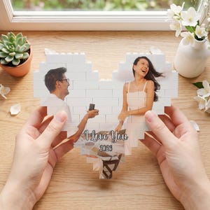 Puede incluir: Un collage de fotos en forma de corazón hecho de bloques blancos, con una pareja. El collage tiene el texto "I Love You" y el año "2026". El collage está sostenido por dos manos, con una mesa de madera y una ventana en el fondo.