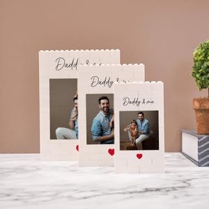 Puede incluir: Tres marcos de fotos blancos con diseño de bloques de construcción. Cada marco muestra una fotografía y las palabras "Daddy" o "Daddy & me" en escritura cursiva. Acentos de corazón rojo debajo.