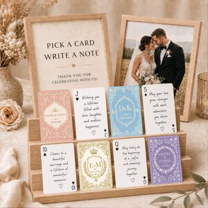 May include: A wooden tiered display stand holding a sign that reads PICK A CARD WRITE A NOTE and a framed photograph of a couple. Several playing cards with custom text and floral designs in pink, blue, gold, and purple are arranged on the stand.