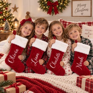 May include: Four red velvet Christmas stockings with white faux fur cuffs, each personalised with white cursive embroidery reading Olivia, Liam, Charlotte, and Noah. The stockings are held by four children sitting on a bed with a white knit blanket.