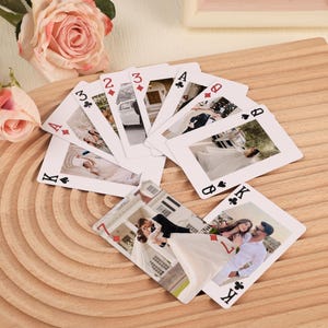 Puede incluir: Un juego de cartas, cada una con una foto de boda diferente. Las cartas están dispuestas en abanico sobre una superficie de madera, con rosas rosas en el fondo. Las cartas muestran diferentes palos y números.
