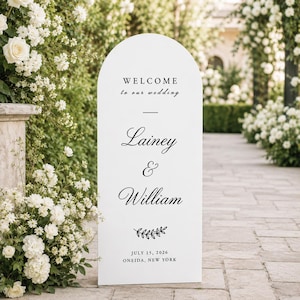 May include: A white arched wedding welcome sign with black text reading WELCOME to our wedding, Lainey & William, July 15, 2026, ONEIDA, NEW YORK, featuring a small decorative leaf illustration centred above the date and location details.