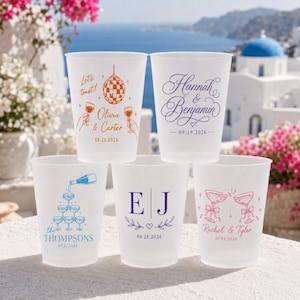 May include: Five frosted plastic cups with custom designs for a wedding or party. The cups feature text and graphics in various colours, including a disco ball, champagne pouring, initials, and names with dates. The background is a scenic view.