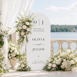 May include: A white arched wedding welcome sign featuring black text that reads O J, WELCOME TO OUR WEDDING, OLIVIA and JULIAN, and JULY 15, 2026 ONEIDA, NEW YORK. The sign is decorated with large arrangements of white roses and greenery.