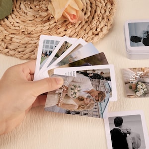 Puede incluir: Una mano sostiene un abanico de tarjetas rectangulares con fotos de boda. Las tarjetas muestran imágenes de novios, con texto en algunas tarjetas. Una pequeña lata blanca con una imagen de vaca está en el fondo.