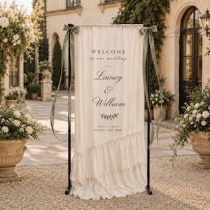 May include: A tall, cream-coloured fabric wedding welcome sign hanging on a black metal frame. The sign features black text reading "WELCOME to our wedding", "Lainey & William", a leaf graphic, and "JULY 15, 2026 ONEIDA, NEW YORK". Sage green ribbons tie the fabric.
