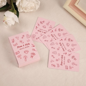 May include: A pink gift box and matching cards are decorated with red line art of celebratory icons. The box and cards display the names "Brian & Kelly" and the date "06.01.2026". The cards are scattered around the box.