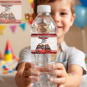 A young boy smiling while holding up a clear water bottle featuring a custom red cartoon racing car label that reads "Mateo is 5". The background shows a blurry kid's birthday party with colorful balloons and party hats. This editable template creates a personalized printable water bottle label for custom race track themed boys birthday party decorations.
