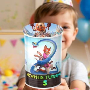 Mockup of a young boy wearing a party hat holding a personalized Zootopia themed Pringles potato chip can. The custom chip wrapper label features Judy Hopps and Nick Wilde cartoon characters with vibrant blue and yellow balloons and birthday decorations blurred in the background.