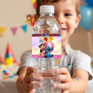 A smiling boy holding a clear water bottle wrapped in a custom Lilo and Stitch birthday party label. The label reads 'Emma is turning 4' with a tropical pink and blue Hawaiian theme. The background is a brightly lit, blurred kids birthday party scene featuring colorful balloons, party hats, and festive pennant banners, showcasing the printable party decor in use.