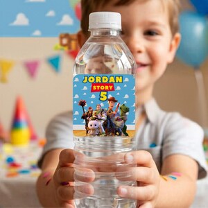 A smiling young boy holding a clear water bottle featuring a personalized Toy Story birthday label. The custom label reads "Jordan Story 5" with Woody, Buzz, and friends. The background shows a colorful, blurred birthday party setting with balloons and party hats. Creative idea for custom kids birthday party supplies, DIY favors, and printable party decorations.