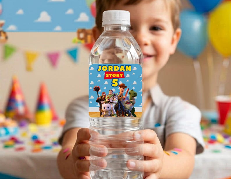 A smiling young boy holding a clear water bottle featuring a personalized Toy Story birthday label. The custom label reads "Jordan Story 5" with Woody, Buzz, and friends. The background shows a colorful, blurred birthday party setting with balloons and party hats. Creative idea for custom kids birthday party supplies, DIY favors, and printable party decorations.