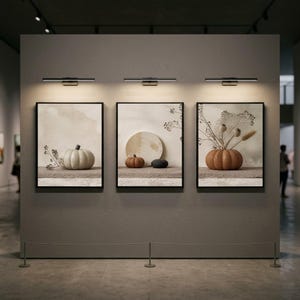 May include: Three rectangular framed wall art prints displayed on a gray wall, each illuminated by a black picture light. The prints feature neutral-toned still life compositions of pumpkins, dried botanical stems, and circular stone shapes.