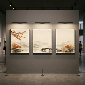 Peut inclure: Trois peintures de paysage encadrées de cadres noirs sont exposées sur un mur gris. Chaque peinture représente une scène sereine avec des montagnes, de l'eau et des arbres dans des tons orange et marron. Au-dessus de chaque peinture se trouve un luminaire noir.