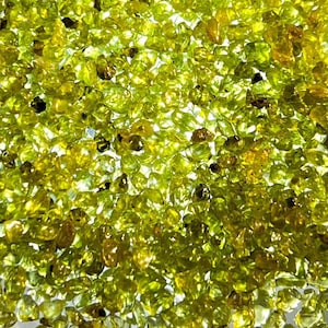 May include: Close-up of a collection of small, irregularly shaped peridot gemstones. The stones are primarily a vibrant, translucent green with hints of yellow and brown, creating a sparkling, textured appearance. The gemstones are scattered across the image.