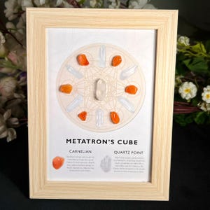 Könnte beinhalten: Ein heller Holzrahmen mit einem Druck von Metatrons Würfel, auf dem vier orangefarbene Karneolsteine und acht klare Quarzkristallspitzen kreisförmig angeordnet sind. Der Text lautet Metatron's Cube, Carnelian und Quartz Point.
