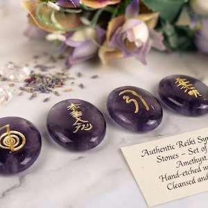 Op de afbeelding: Een set van vier gladde, ovaalvormige amethiststenen, elk voorzien van een met de hand in goud gegraveerd Reiki-symbool. Een klein wit kaartje vergezelt de stenen, bedrukt met de tekst: Authentic Reiki Symbol Stones - Set of 4. Amethyst. Hand-etched with Gold. Cleansed and Charged.